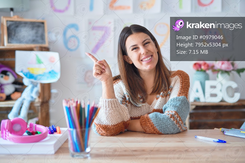 Young beautiful teacher woman wearing sweater and glasses sitting on desk at kindergarten with a big smile on face, pointing with hand and finger to the side looking at the camera.