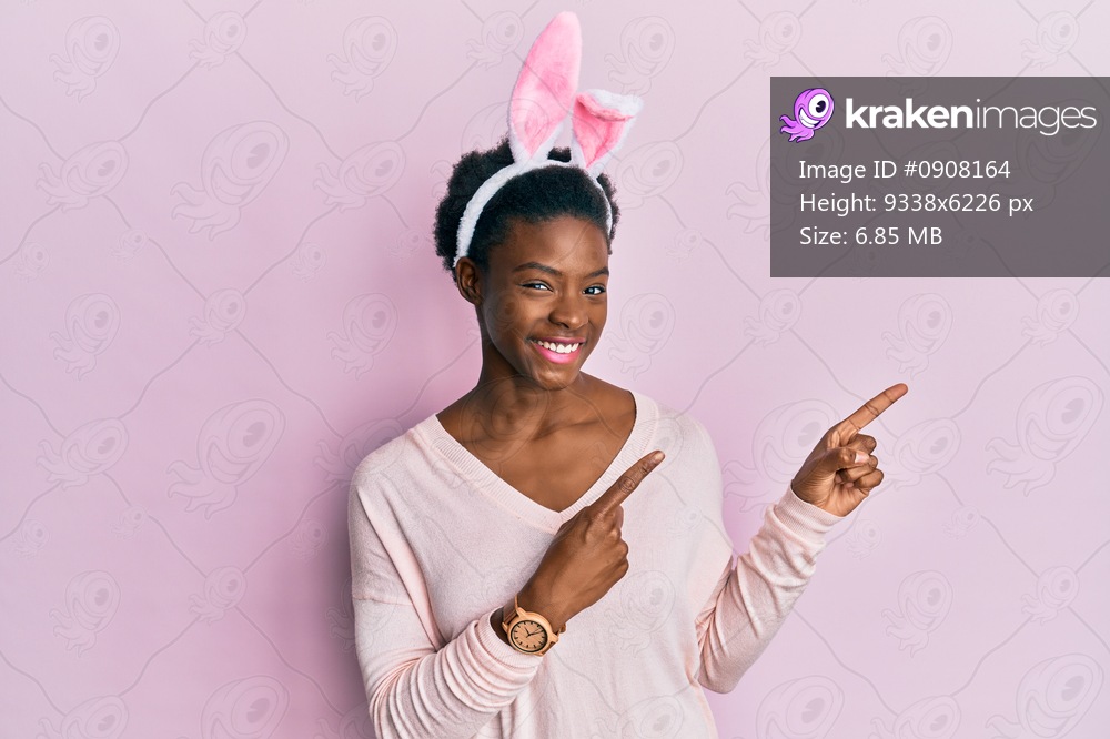 Young african american girl wearing cute easter bunny ears smiling and looking at the camera pointing with two hands and fingers to the side. 