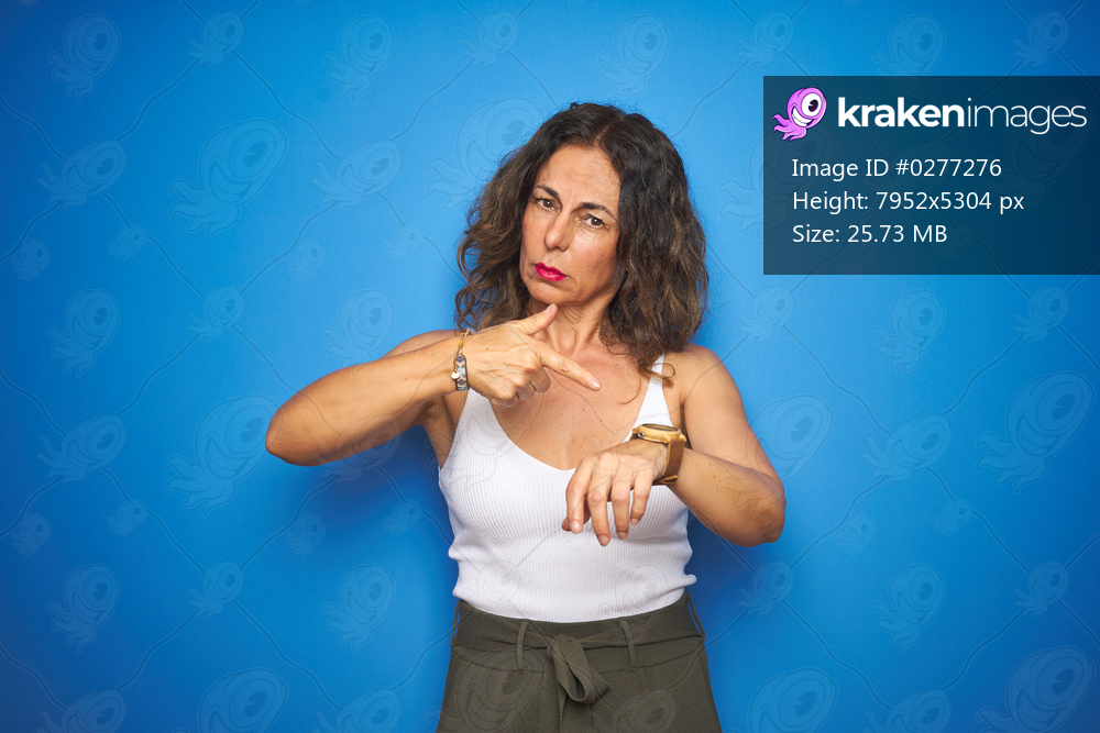 Middle age senior woman with curly hair standing over blue isolated background In hurry pointing to watch time, impatience, upset and angry for deadline delay