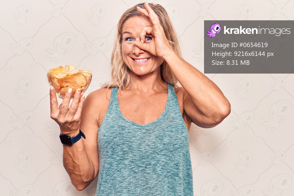 Middle age caucasian blonde woman holding potato chips smiling happy doing ok sign with hand on eye looking through fingers 