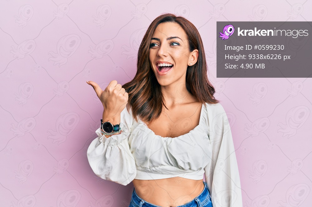 Young brunette woman wearing casual clothes over pink pointing thumb up to the side smiling happy with open mouth 