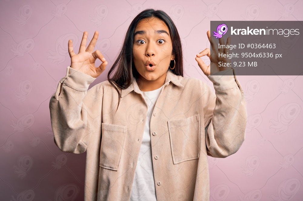 Young beautiful asian woman wearing casual shirt standing over pink background looking surprised and shocked doing ok approval symbol with fingers. Crazy expression
