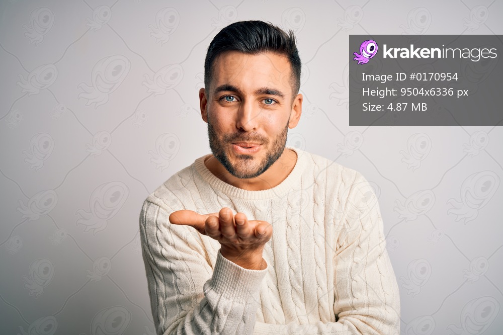Young handsome man wearing casual sweater standing over isolated white background looking at the camera blowing a kiss with hand on air being lovely and sexy. Love expression.