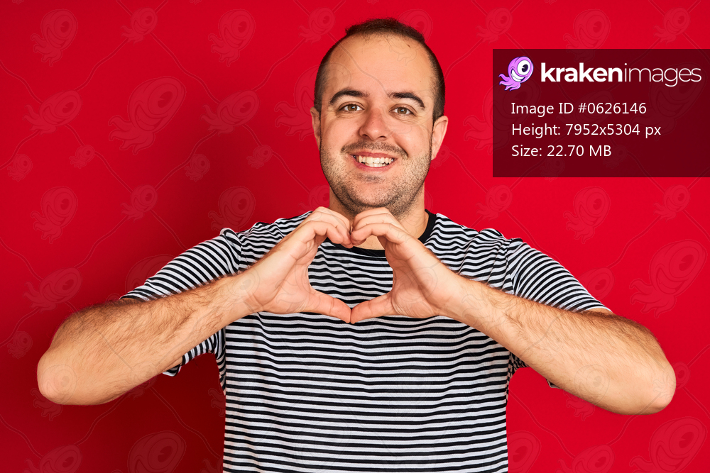 Young man wearing striped navy t-shirt standing over isolated red background smiling in love showing heart symbol and shape with hands. Romantic concept.