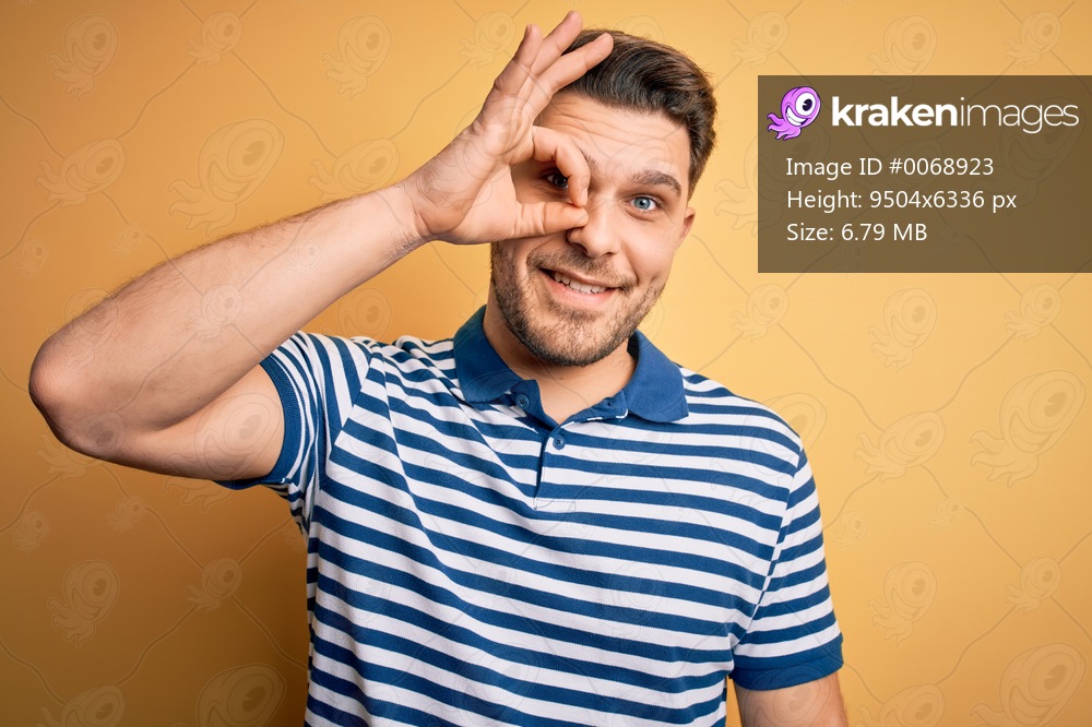 Young man with blue eyes wearing casual striped t-shirt over yellow background doing ok gesture with hand smiling, eye looking through fingers with happy face.