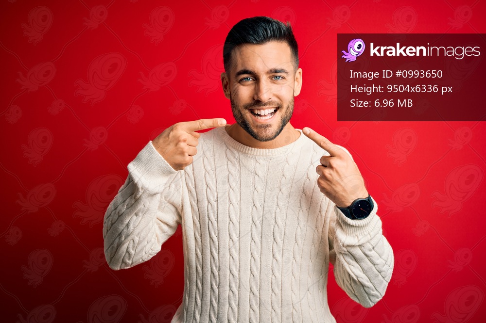 Young handsome man wearing casual white sweater standing over isolated red background smiling cheerful showing and pointing with fingers teeth and mouth. Dental health concept.