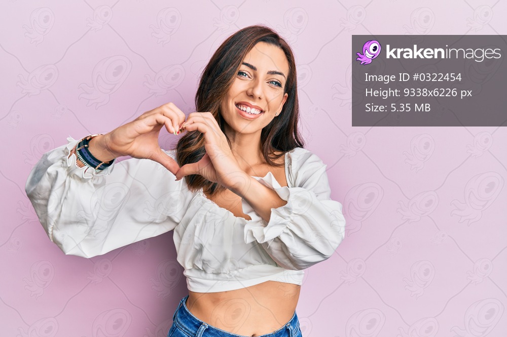Young brunette woman wearing casual clothes over pink smiling in love doing heart symbol shape with hands. romantic concept. 
