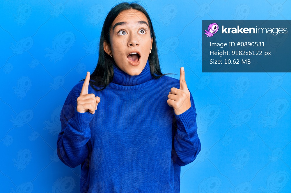 Young asian woman wearing casual winter sweater amazed and surprised looking up and pointing with fingers and raised arms. 