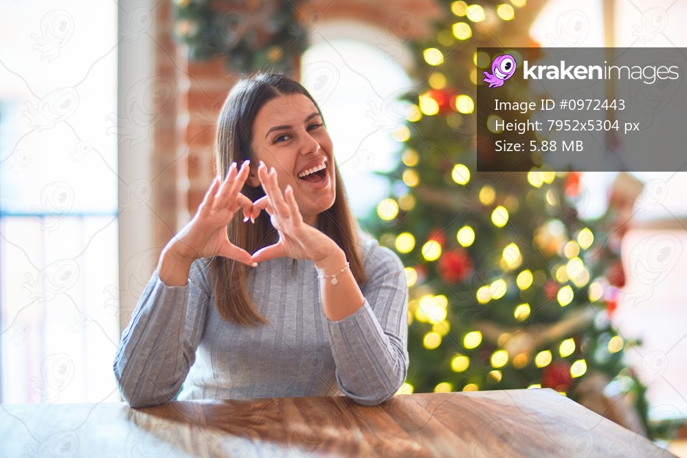 Young beautiful woman sitting at the table at home around christmas tree and decoration smiling in love showing heart symbol and shape with hands. Romantic concept.