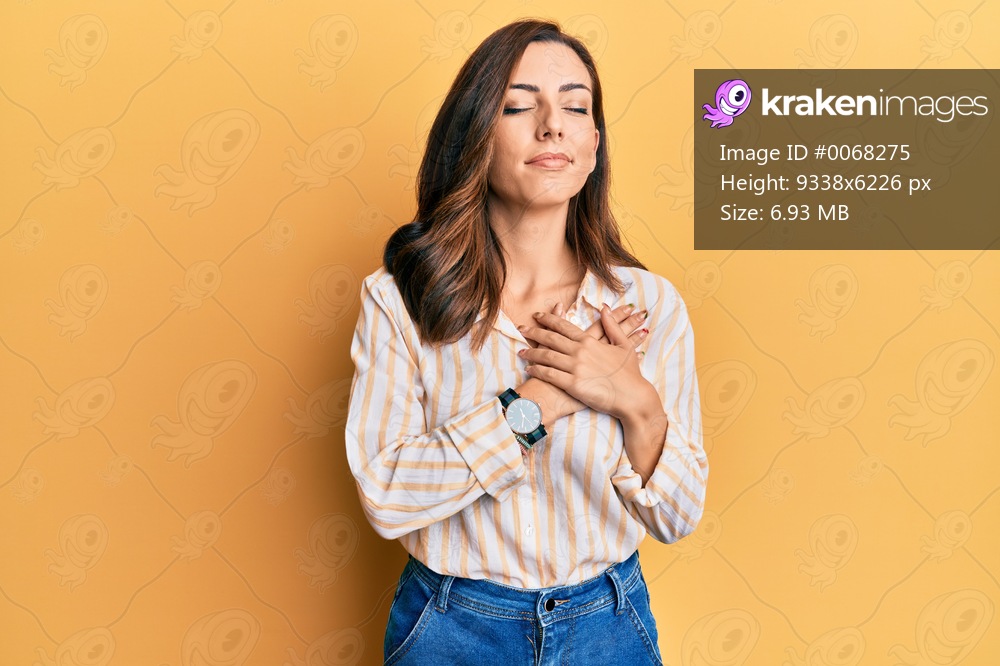 Young brunette woman wearing casual clothes over yellow background smiling with hands on chest, eyes closed with grateful gesture on face. health concept. 