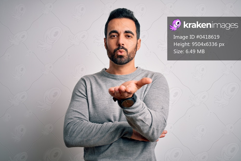 Young handsome man with beard wearing casual sweater standing over white background looking at the camera blowing a kiss with hand on air being lovely and sexy. Love expression.