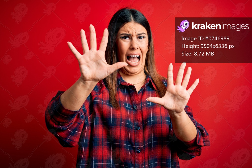 Young beautiful woman wearing casual shirt over red background afraid and terrified with fear expression stop gesture with hands, shouting in shock. Panic concept.
