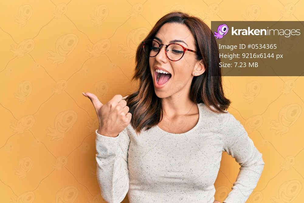 Young brunette woman wearing casual clothes and glasses pointing thumb up to the side smiling happy with open mouth 