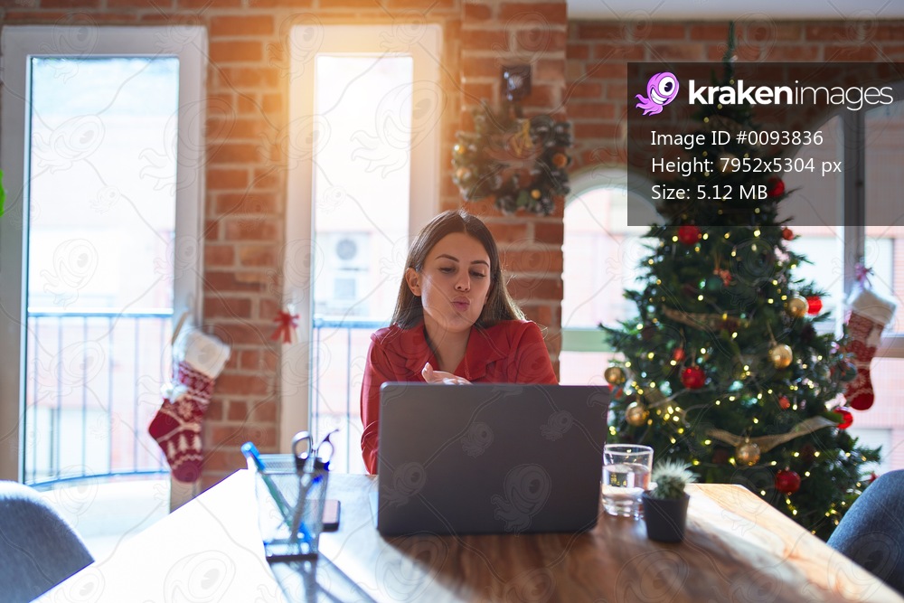 Beautiful woman sitting at the table working with laptop at home around christmas tree looking at the camera blowing a kiss with hand on air being lovely and sexy. Love expression.