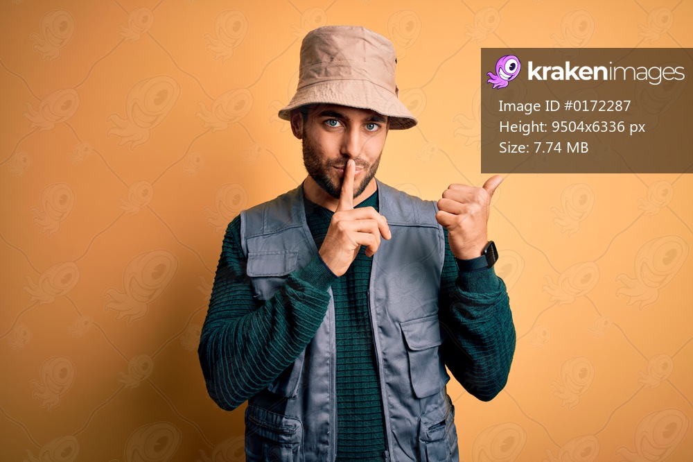 Handsome tourist man with beard on vacation wearing explorer hat over yellow background asking to be quiet with finger on lips pointing with hand to the side. Silence and secret concept.