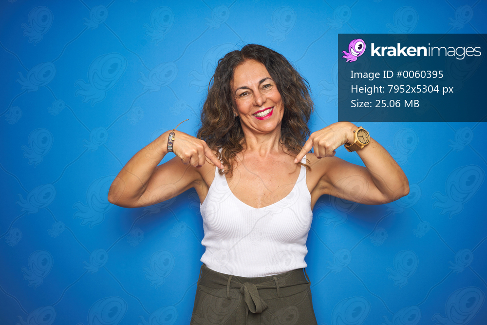 Middle age senior woman with curly hair standing over blue isolated background looking confident with smile on face, pointing oneself with fingers proud and happy.