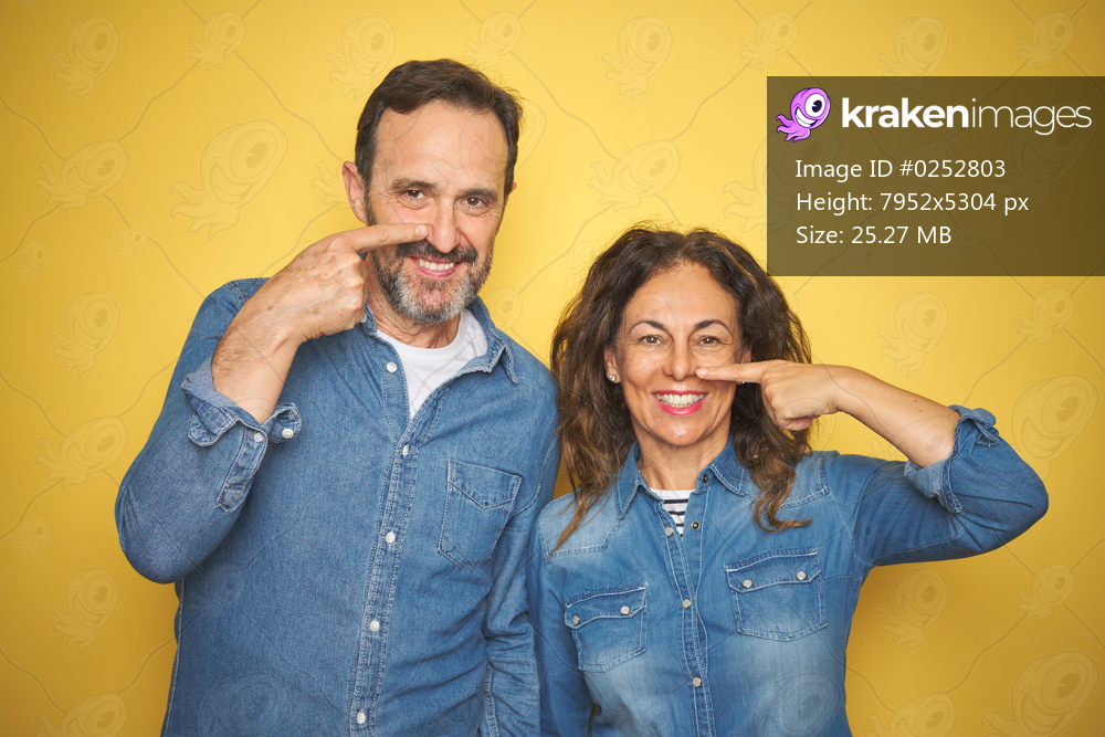 Beautiful middle age couple together standing over isolated yellow background Pointing with hand finger to face and nose, smiling cheerful. Beauty concept