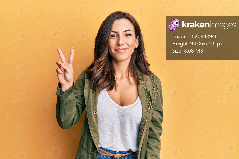 Young brunette woman wearing casual clothes over yellow background smiling with happy face winking at the camera doing victory sign. number two. 