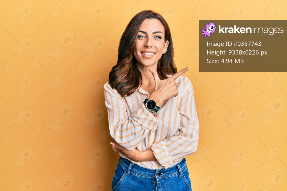 Young brunette woman wearing casual clothes over yellow background smiling cheerful pointing with hand and finger up to the side 