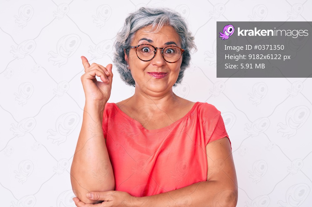 Senior hispanic grey- haired woman wearing casual clothes and glasses with a big smile on face, pointing with hand and finger to the side looking at the camera. 