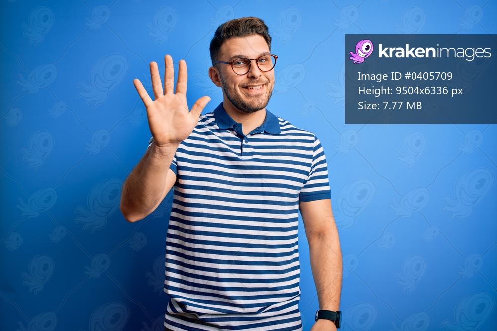 Young man with blue eyes wearing glasses and casual striped t-shirt over blue background showing and pointing up with fingers number five while smiling confident and happy.