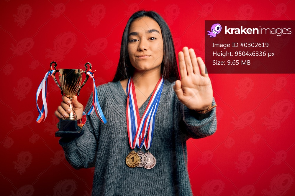 Young beautiful champion asian woman holding trophy wearing medals over red background with open hand doing stop sign with serious and confident expression, defense gesture