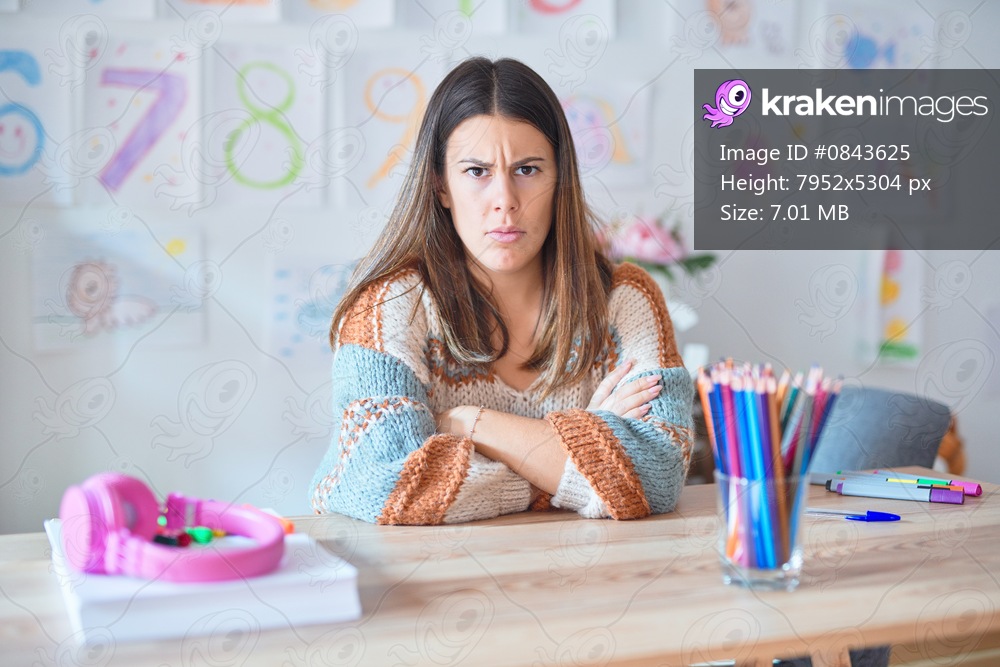 Young beautiful teacher woman wearing sweater and glasses sitting on desk at kindergarten skeptic and nervous, disapproving expression on face with crossed arms. Negative person.