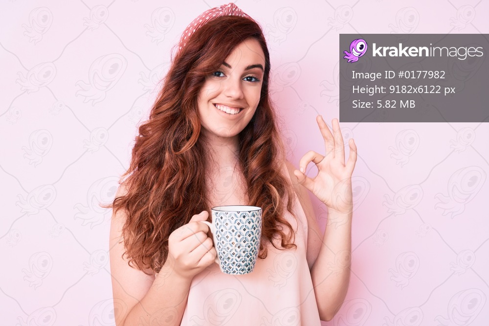 Young beautiful woman holding coffee doing ok sign with fingers, smiling friendly gesturing excellent symbol 