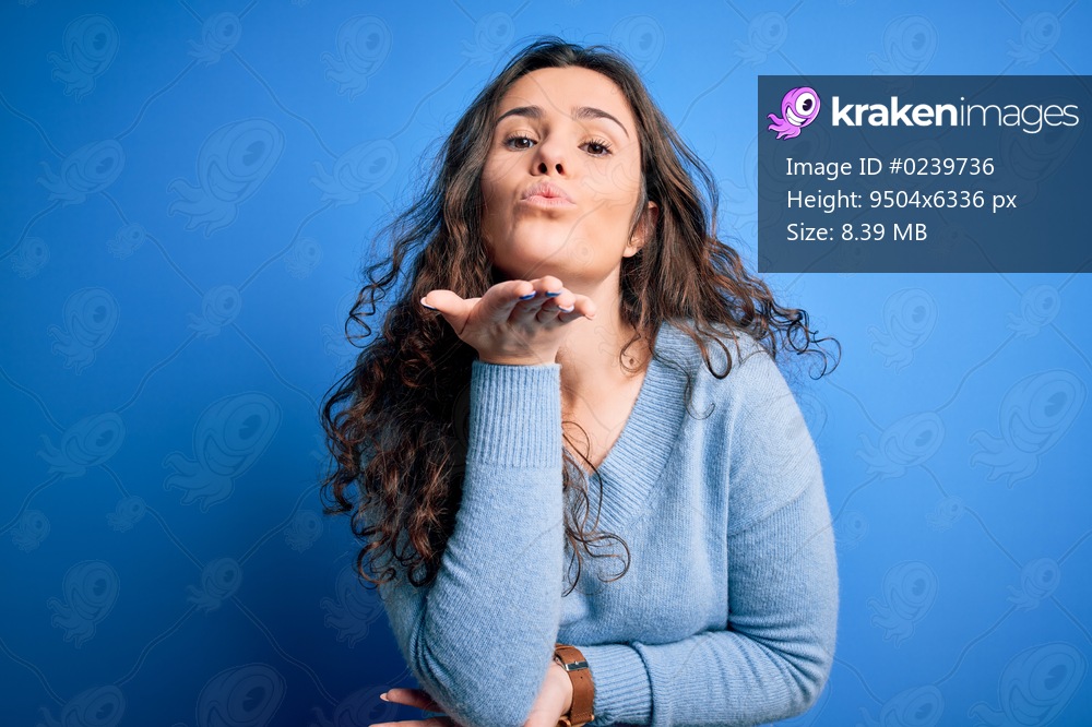 Young beautiful woman with curly hair wearing blue casual sweater over isolated background looking at the camera blowing a kiss with hand on air being lovely and sexy. Love expression.