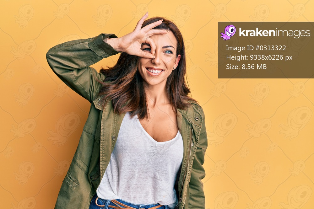 Young brunette woman wearing casual clothes over yellow background smiling happy doing ok sign with hand on eye looking through fingers 