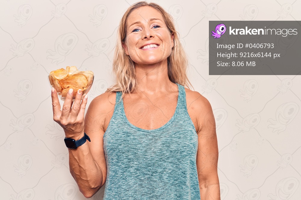 Middle age caucasian blonde woman holding potato chips looking positive and happy standing and smiling with a confident smile showing teeth 
