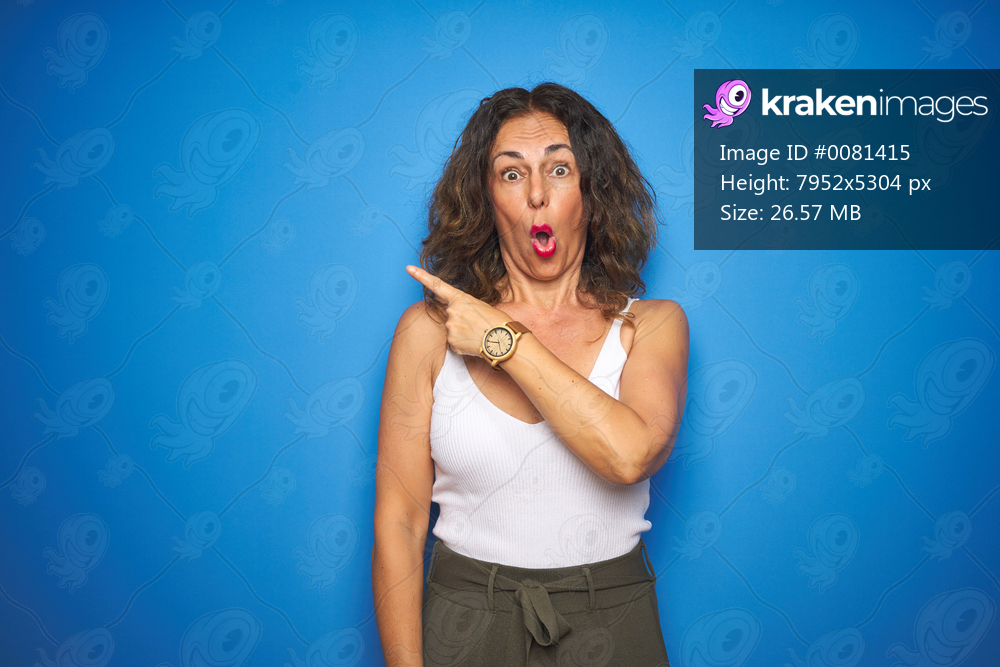 Middle age senior woman with curly hair standing over blue isolated background Surprised pointing with finger to the side, open mouth amazed expression.