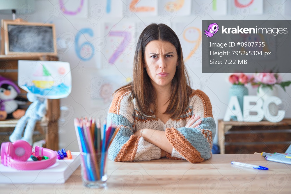 Young beautiful teacher woman wearing sweater and glasses sitting on desk at kindergarten skeptic and nervous, disapproving expression on face with crossed arms. Negative person.
