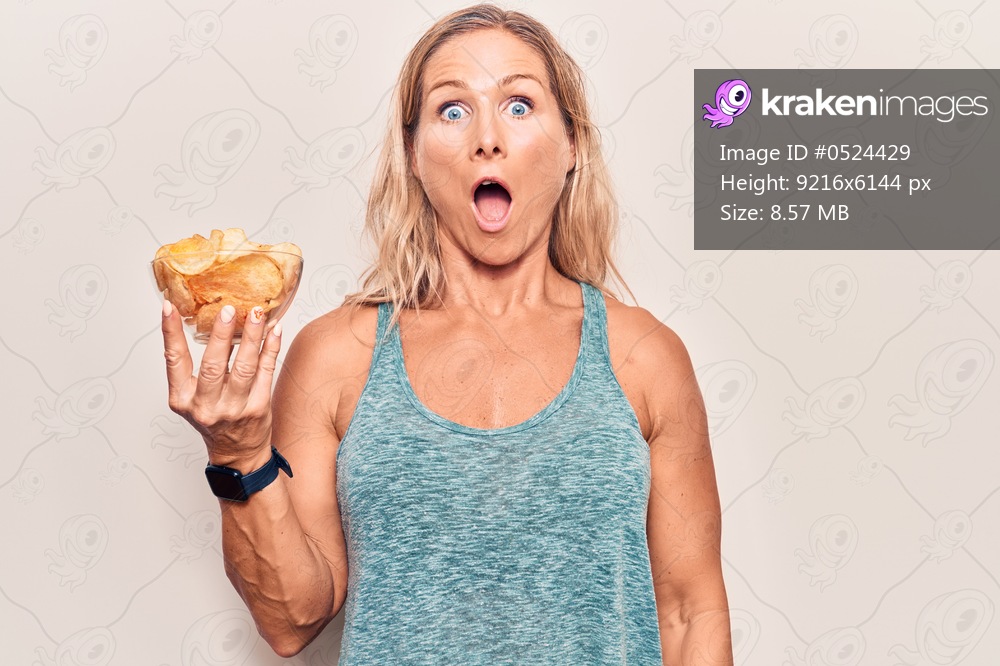 Middle age caucasian blonde woman holding potato chips scared and amazed with open mouth for surprise, disbelief face 