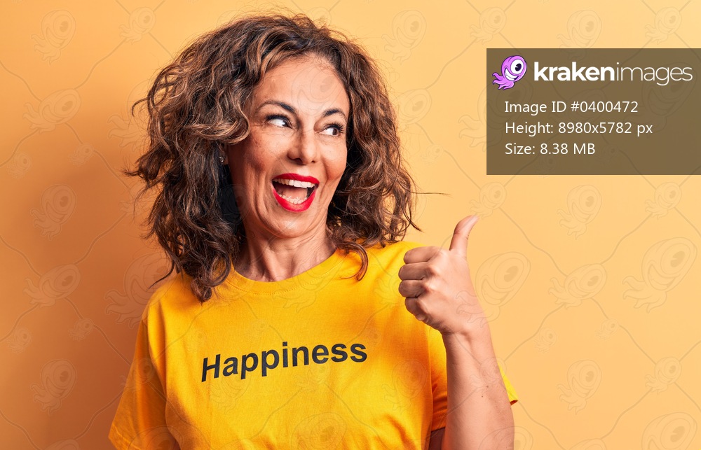Middle age beautiful woman wearing t-shirt with happiness word over yellow background pointing thumb up to the side smiling happy with open mouth