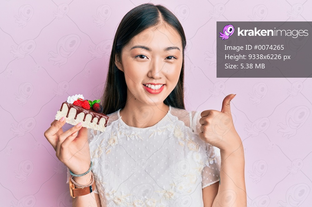 Young chinese woman eating a tasty cake smiling happy and positive, thumb up doing excellent and approval sign 