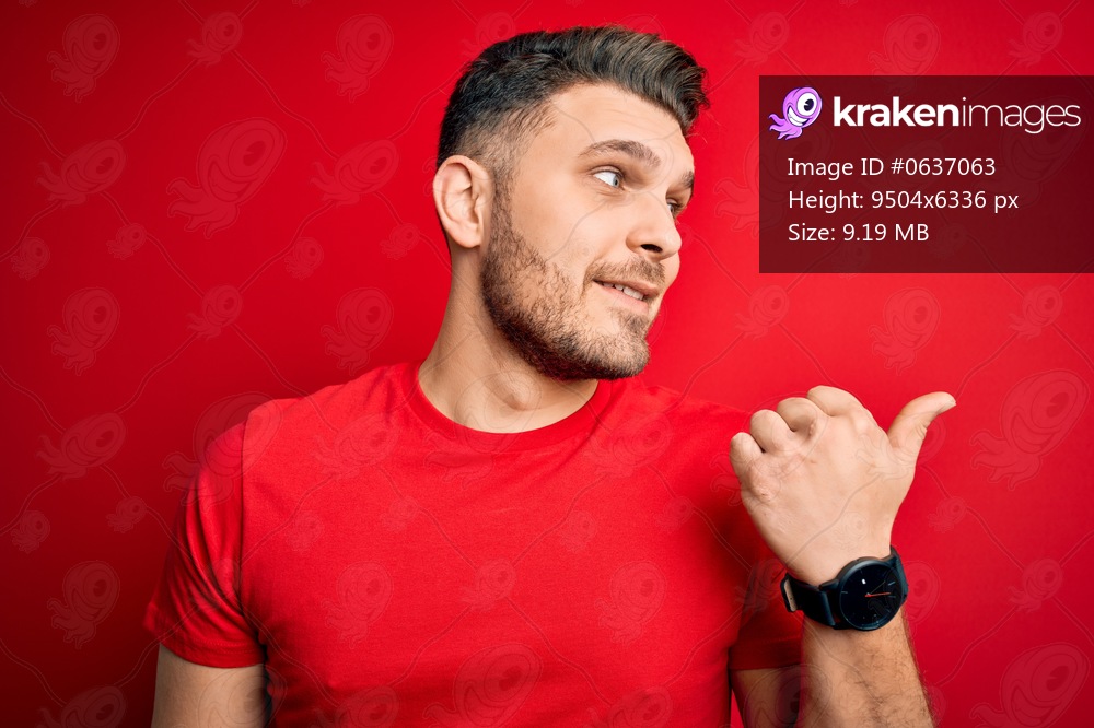 Young man with blue eyes wearing casual t-shirt over red isolated background smiling with happy face looking and pointing to the side with thumb up.