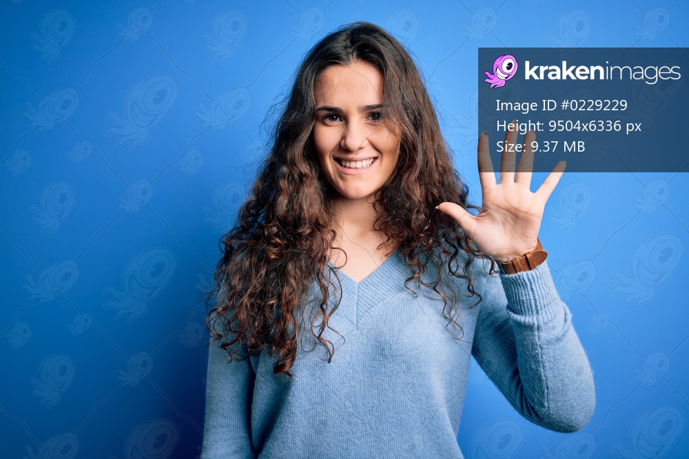 Young beautiful woman with curly hair wearing blue casual sweater over isolated background showing and pointing up with fingers number five while smiling confident and happy.