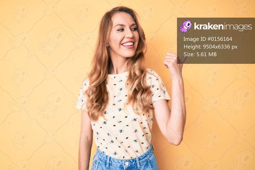 Young caucasian woman with blond hair wearing casual summer clothes pointing thumb up to the side smiling happy with open mouth 