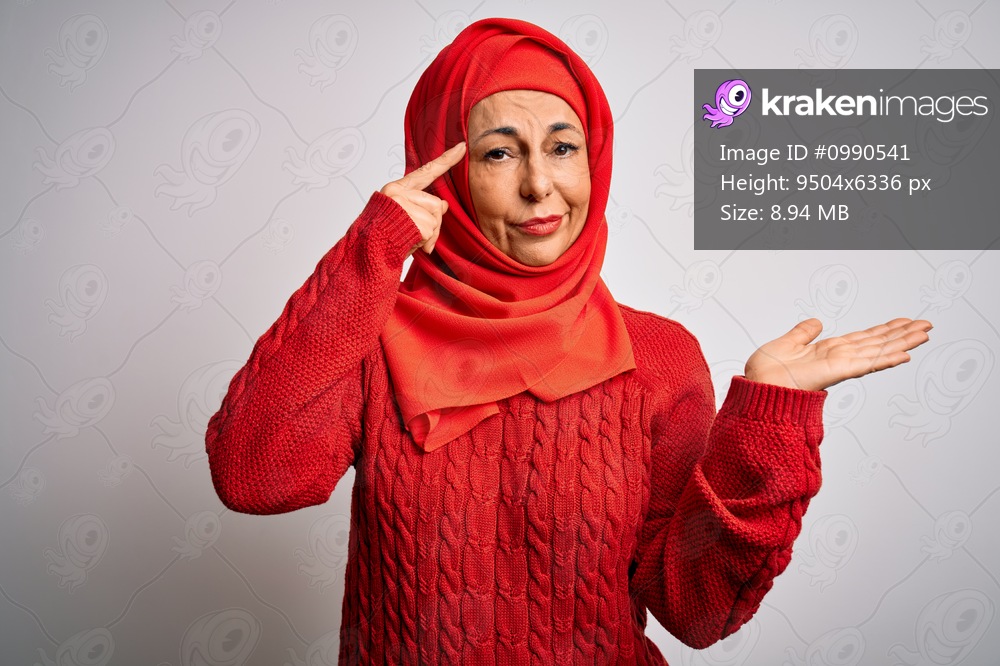 Middle age brunette woman wearing muslim traditional hijab over isolated white background confused and annoyed with open palm showing copy space and pointing finger to forehead. Think about it.