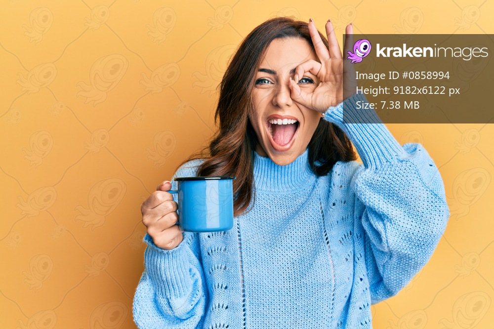 Young brunette woman holding a cup of coffee smiling happy doing ok sign with hand on eye looking through fingers 