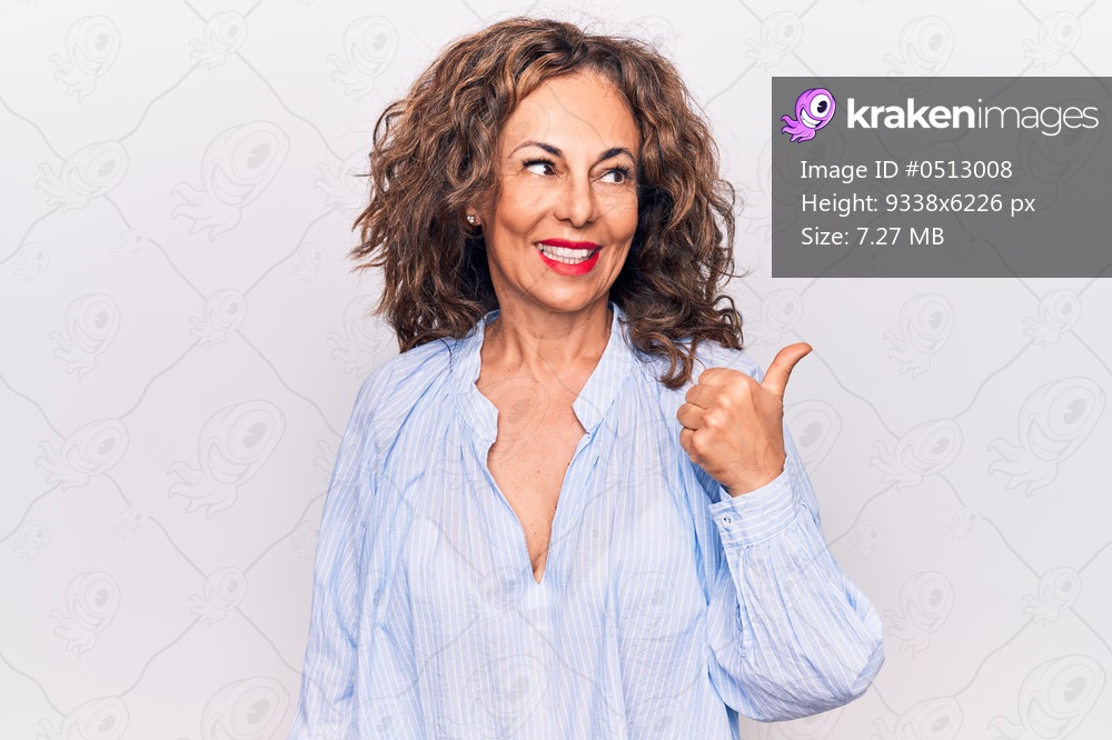 Middle age beautiful brunette woman wearing striped shirt standing over white background pointing thumb up to the side smiling happy with open mouth