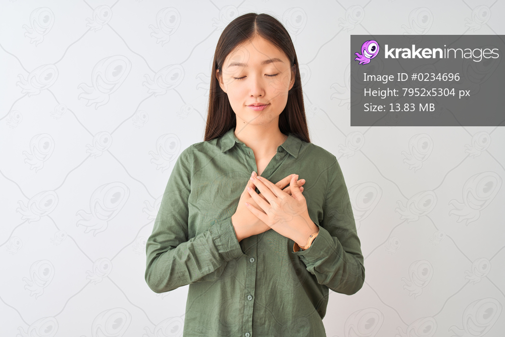 Young chinese woman wearing casual green shirt standing over isolated white background smiling with hands on chest with closed eyes and grateful gesture on face. Health concept.
