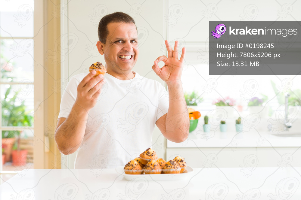 Middle age man eating chocolate chip muffins at home doing ok sign with fingers, excellent symbol