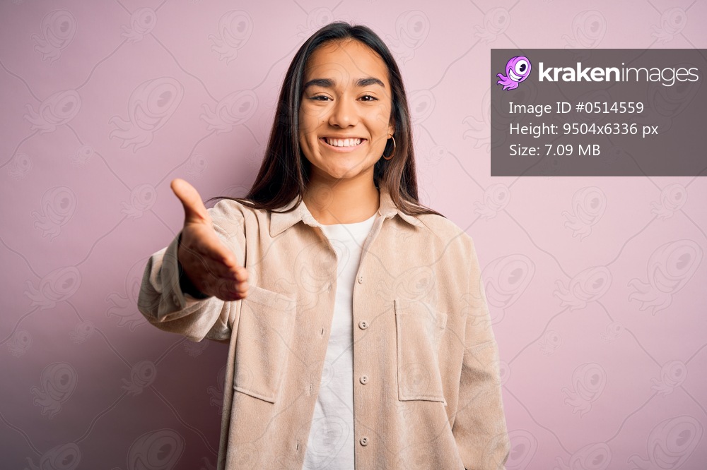 Young beautiful asian woman wearing casual shirt standing over pink background smiling friendly offering handshake as greeting and welcoming. Successful business.