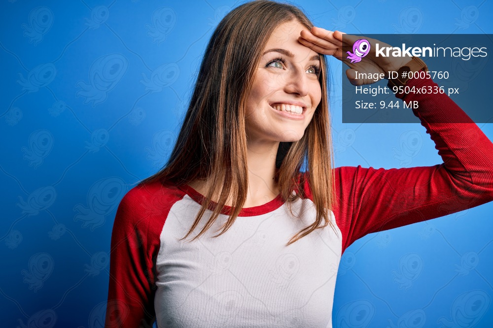 Young beautiful redhead woman wearing casual t-shirt over isolated blue background very happy and smiling looking far away with hand over head. Searching concept.
