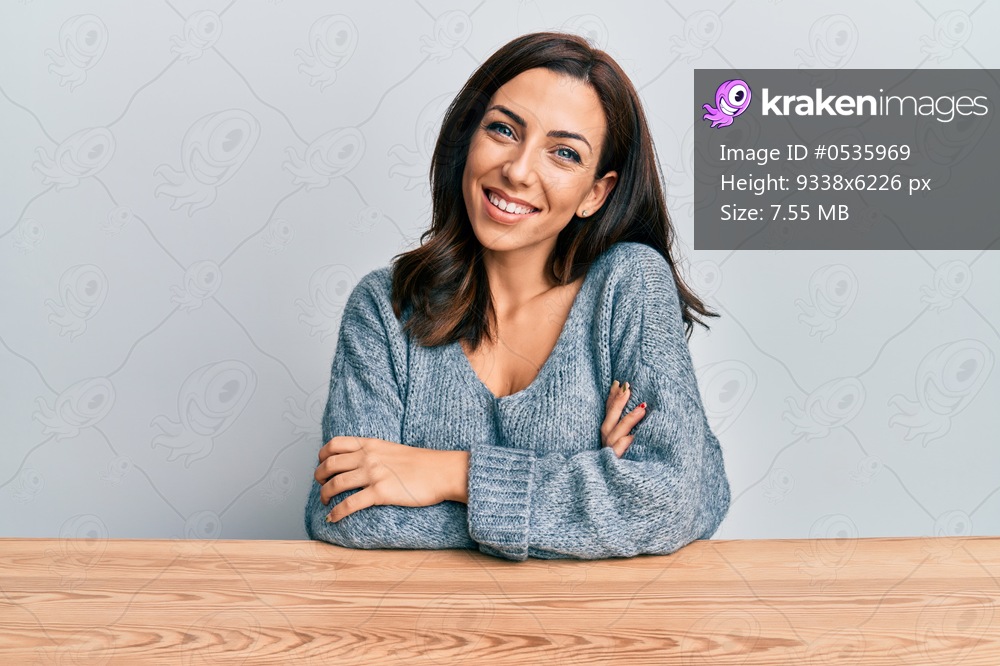 Young brunette woman wearing casual sweater sitting on the table happy face smiling with crossed arms looking at the camera. positive person. 