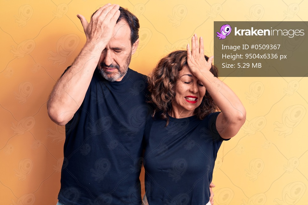 Middle age beautiful couple wearing t-shirt standing over isolated yellow background surprised with hand on head for mistake, remember error. Forgot, bad memory concept.