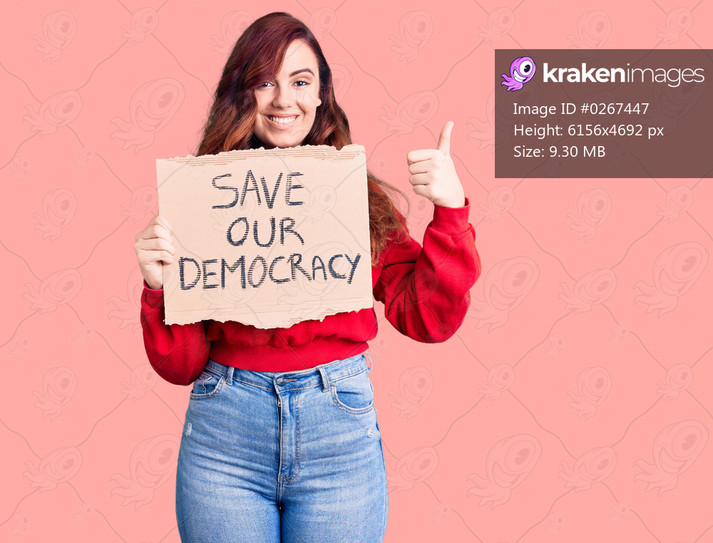 Young beautiful woman holding save our democracy protest banner smiling happy and positive, thumb up doing excellent and approval sign 