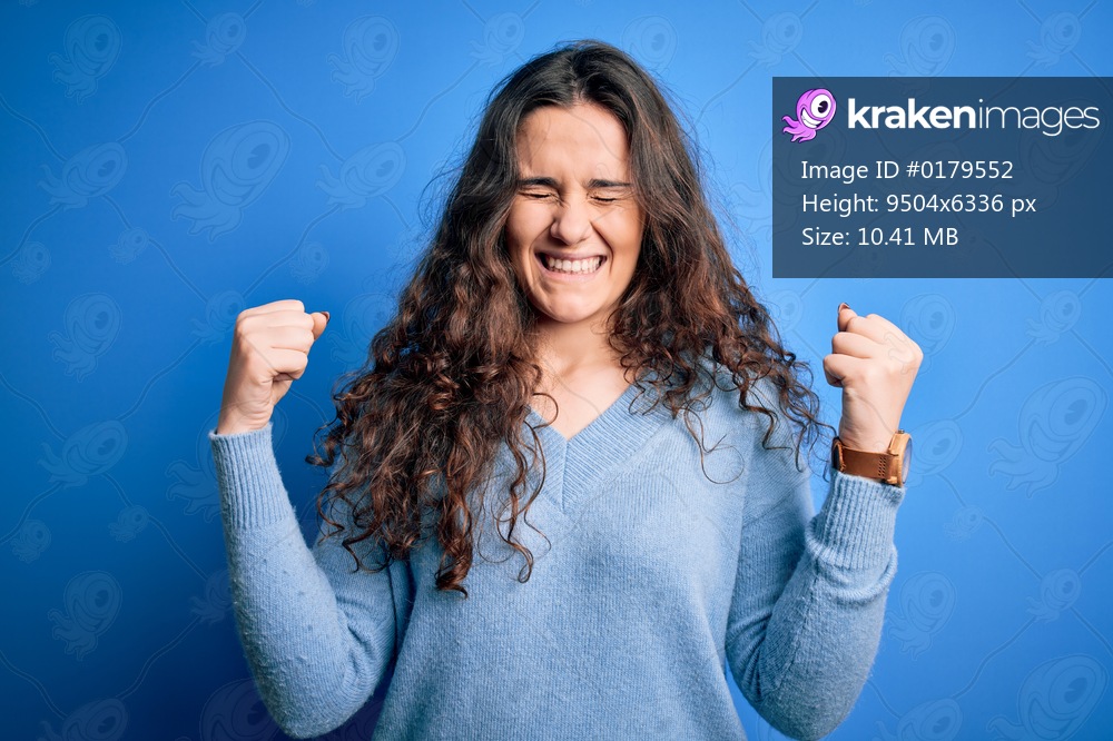 Young beautiful woman with curly hair wearing blue casual sweater over isolated background very happy and excited doing winner gesture with arms raised, smiling and screaming for success. Celebration concept.